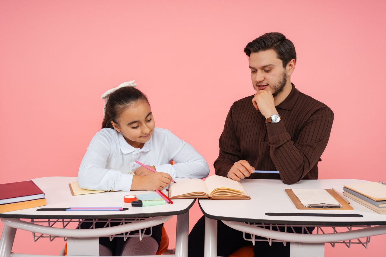 À Propos A teacher helps a young student with writing tasks at a desk indoors.