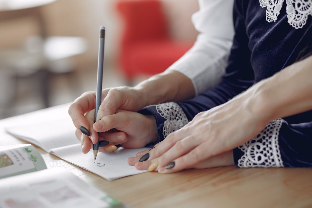 Nos Formules High angle of crop unrecognizable black female teacher explaining task to focused little schoolboy sitting at desk near attentive Asian classmate writing in notebook