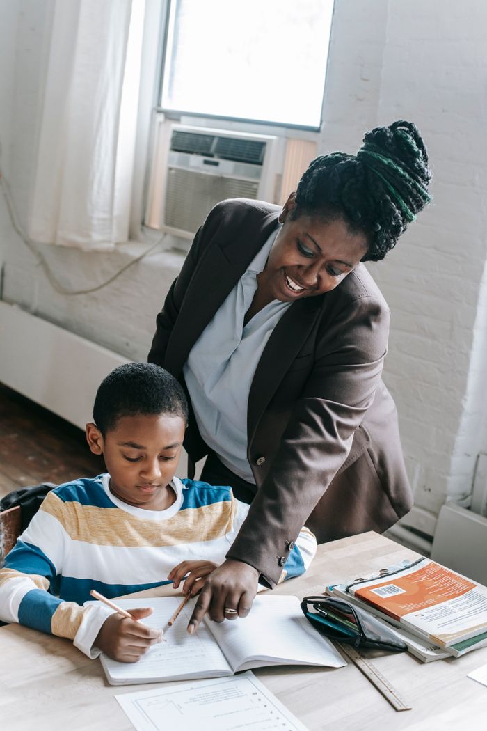 Accueil High angle of positive adult African American female teacher in formal clothes helping little boy doing assignment during lesson in school